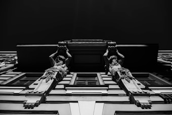 Two sculpted figures in an architectural design are viewed from below, extending upwards on a classical façade. The figures appear to be part of decorative column-like structures, showcasing detailed craftsmanship and ornate features.