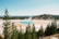 A vibrant geological hot spring surrounded by lush green trees and distant hills under a clear sky. The hot spring features vivid blue and orange hues, with steam rising from its surface, creating a contrasting natural spectacle.
