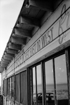 A weathered wooden building with an old sign on the side that reads 'BEST EAST OR WEST', featuring a row of hanging light bulbs under the eaves. Large windows reflect a glimpse of an ocean vista with waves in the background.