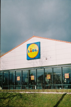 A large, single-story supermarket building with a prominent glass facade. The structure features a white exterior with a sloped, terracotta-colored roof. Near the top of the building, a brightly colored sign with a blue border and a yellow circle is prominently displayed. Below, the windows showcase promotional posters inside.