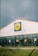 A large, single-story supermarket building with a prominent glass facade. The structure features a white exterior with a sloped, terracotta-colored roof. Near the top of the building, a brightly colored sign with a blue border and a yellow circle is prominently displayed. Below, the windows showcase promotional posters inside.
