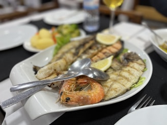A white platter is filled with a variety of grilled seafood including a prawn, fish fillets, and a lemon wedge. The seafood is accompanied by a mix of salad on the side. Cutlery is placed on the platter, and there are other dishes, including a blurred plate of potatoes and a glass of white wine, visible in the background.