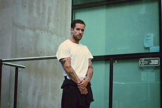 A man with tattoos on his arms is standing, wearing a white t-shirt and dark pants. He is positioned near a glass wall with a sign in Spanish. The setting appears to be urban and modern, featuring a concrete wall and metal railings.