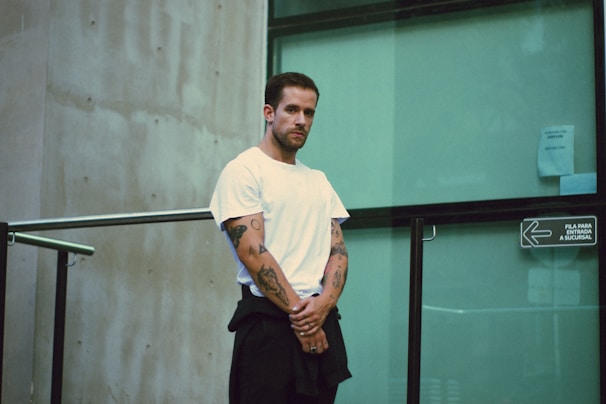 A man with tattoos on his arms is standing, wearing a white t-shirt and dark pants. He is positioned near a glass wall with a sign in Spanish. The setting appears to be urban and modern, featuring a concrete wall and metal railings.
