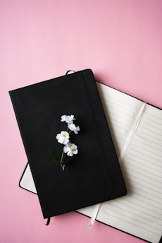 A black closed notebook with white flowers placed on top sits on an open lined notebook against a pink background. The open notebook has cream-colored pages and a white ribbon bookmark.