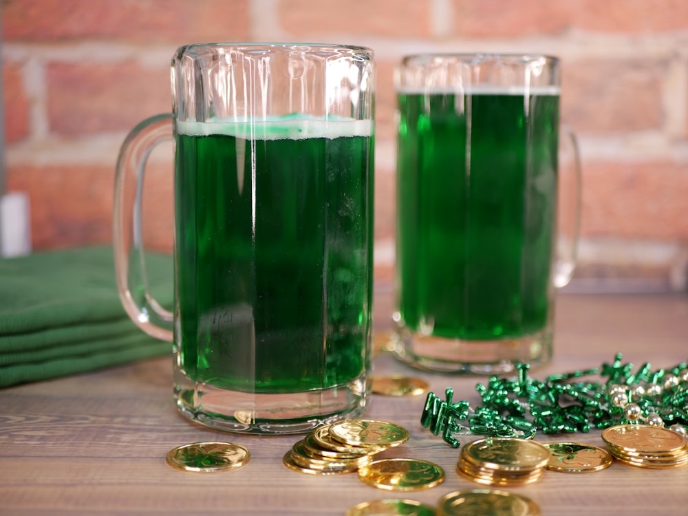Dos tazas llenas de líquido verde junto a monedas de oro