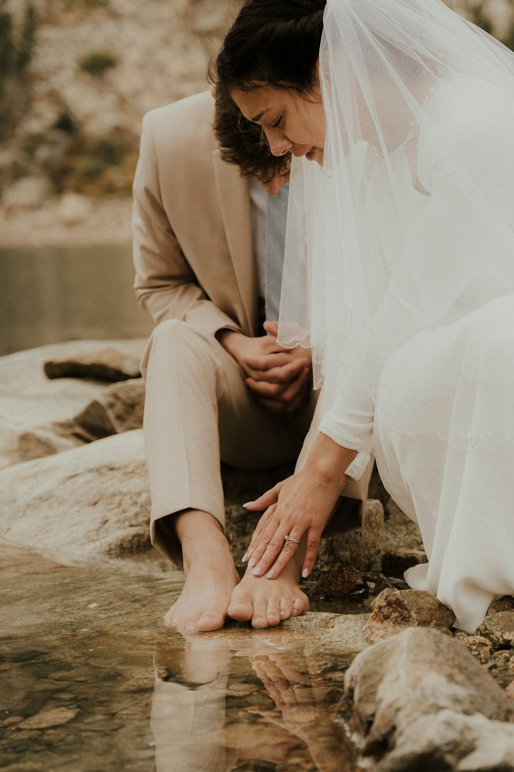 a bride and groom sitting on rocks by the water