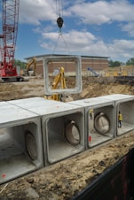 A construction site featuring a large red crane lifting a concrete section with a massive hole, positioned above a series of similar concrete sections placed in a trench. Workers wearing helmets and high-visibility vests are visible among the structures. In the background, there is a red and brown building and a clear blue sky with some clouds.