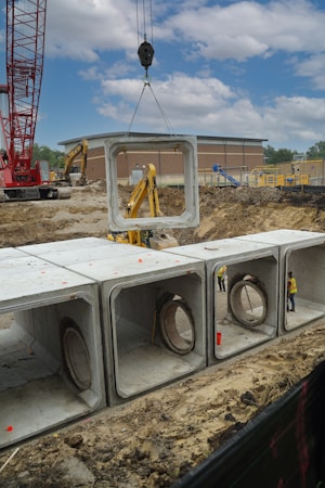 A construction site featuring a large red crane lifting a concrete section with a massive hole, positioned above a series of similar concrete sections placed in a trench. Workers wearing helmets and high-visibility vests are visible among the structures. In the background, there is a red and brown building and a clear blue sky with some clouds.