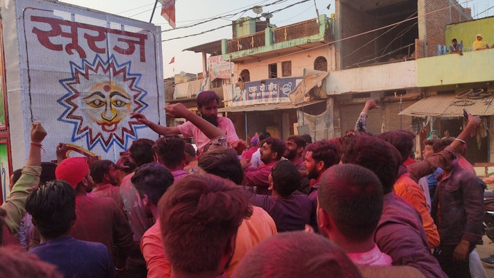 A large group of people, mostly men, gathered on a street, colored in hues of red and pink likely due to a festival. They are standing near a colorful banner featuring a sun-like face with Hindi text on it. The scene is filled with energy and excitement as the crowd seems to be participating in a celebration. The background shows a street with buildings, some shops, and houses.