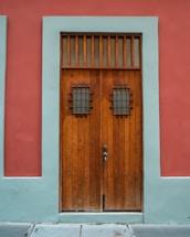 A wooden door with vertical panels surrounded by a teal border set against a red wall. The door features two small windows with metal grilles and a vintage-style handle.