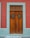 A wooden door with vertical panels surrounded by a teal border set against a red wall. The door features two small windows with metal grilles and a vintage-style handle.