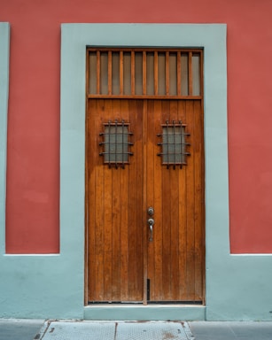 A wooden door with vertical panels surrounded by a teal border set against a red wall. The door features two small windows with metal grilles and a vintage-style handle.