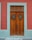 A wooden door with vertical panels surrounded by a teal border set against a red wall. The door features two small windows with metal grilles and a vintage-style handle.