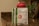A packet of vegan protein powder is displayed on a kitchen countertop next to a red shaker bottle. The label on the packet reads 'Sprouts Vegan Protein, Salted Caramel flavored' and specifies features like easy to digest, gluten-free, and soy-free. The background is a blurred kitchen setting with dim lighting and a hint of a green plant.