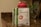 A packet of vegan protein powder is displayed on a kitchen countertop next to a red shaker bottle. The label on the packet reads 'Sprouts Vegan Protein, Salted Caramel flavored' and specifies features like easy to digest, gluten-free, and soy-free. The background is a blurred kitchen setting with dim lighting and a hint of a green plant.