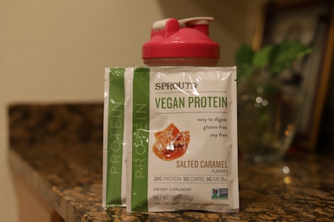 A packet of vegan protein powder is displayed on a kitchen countertop next to a red shaker bottle. The label on the packet reads 'Sprouts Vegan Protein, Salted Caramel flavored' and specifies features like easy to digest, gluten-free, and soy-free. The background is a blurred kitchen setting with dim lighting and a hint of a green plant.