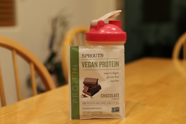 A packet of Sprouts vegan protein powder in chocolate flavor sits on a wooden table. Beside it, there is a red and white shaker bottle. The protein packet highlights features such as being easy to digest, gluten-free, and soy-free.