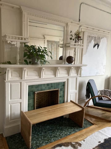 A beautifully decorated interior space featuring a white wooden mantel with decorative columns and moldings. On the mantel, there are lush green plants in a pot and an arrangement of flowers in a vase. A large mirror is mounted above, reflecting part of the room. Below the mantel is a green tiled area suggesting a fireplace with a decorative metal front. In front of the fireplace is a wooden bench on a green woven rug. To the right, there is a stylish armchair with a patterned fabric. The walls are adorned with a piece of abstract art.
