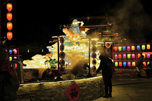 A vibrant nighttime scene at a festival with illuminated lanterns of various colors hanging in a background display. A food stall with steaming pots is in the foreground, and people are standing nearby. A large decorative float features a tiger and bright clouds, casting colorful lights.