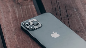 A sleek smartphone with a triple-lens camera rests on a dark wooden surface. Its reflective glass back and the iconic apple logo are prominently visible. The image captures the phone at an angle, highlighting the design and details of the camera module.