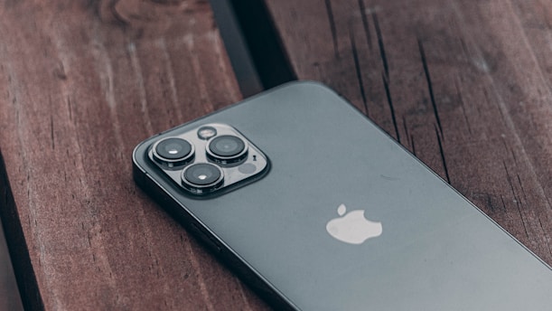 A sleek smartphone with a triple-lens camera rests on a dark wooden surface. Its reflective glass back and the iconic apple logo are prominently visible. The image captures the phone at an angle, highlighting the design and details of the camera module.