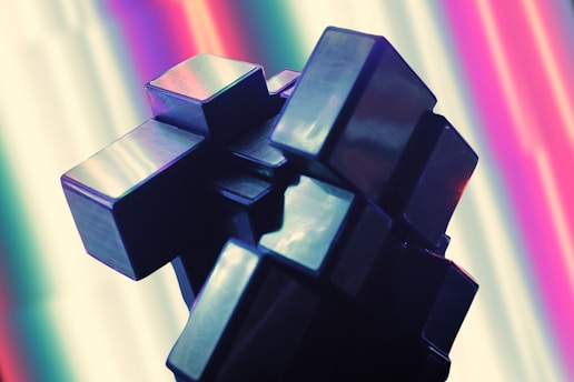 A complex geometric sculpture composed of metallic cubes stacked and interconnected at various angles. The background features a gradient of bright, vivid colors including pink, purple, and green, creating a modern and vibrant atmosphere.