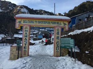 A snow-covered entrance sign welcomes visitors to Ghorepani Poon Hill, with the word 'Namaste' written vertically on pillars. People are seen walking in the snow, and there are wooden and stone structures around. The scene is set in a mountainous area with trees and distant buildings visible.