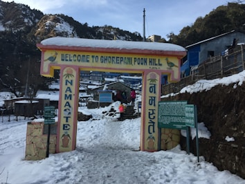 A snow-covered entrance sign welcomes visitors to Ghorepani Poon Hill, with the word 'Namaste' written vertically on pillars. People are seen walking in the snow, and there are wooden and stone structures around. The scene is set in a mountainous area with trees and distant buildings visible.
