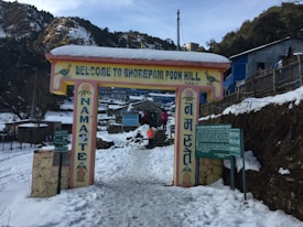 A snow-covered entrance sign welcomes visitors to Ghorepani Poon Hill, with the word 'Namaste' written vertically on pillars. People are seen walking in the snow, and there are wooden and stone structures around. The scene is set in a mountainous area with trees and distant buildings visible.