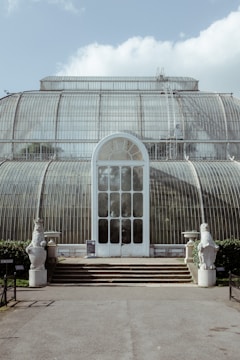 A large, glass greenhouse structure with arched windows and a domed roof. The building has a central entrance with a white door featuring geometric patterns in the glass. Stone steps lead up to the entrance, and two stone statues flank the doorway. The greenhouse is set against a backdrop of a partly cloudy sky, casting soft light on the building.