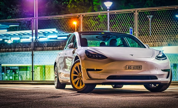 A white Tesla car is parked in an urban setting during nighttime, illuminated by streetlights. The background features a metal fence and a parking area, with a mix of neon and orange lighting.