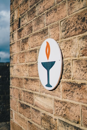 A brick wall features an oval sign with a stylized flame design above a chalice. The sign is colorful and contrasts against the earthy tones of the bricks.