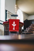 A wooden podium with a red cloth draped over it, featuring a stylized white flame symbol. The background includes rows of pews and an arched window allowing natural light to fill the space.