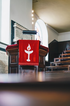 A wooden podium with a red cloth draped over it, featuring a stylized white flame symbol. The background includes rows of pews and an arched window allowing natural light to fill the space.