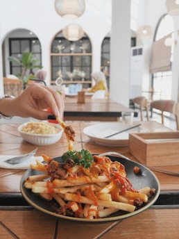 A plate of loaded fries topped with melted cheese, ground meat, and garnished with parsley. A hand is reaching for one of the fries. The setting is a modern and stylish cafe with an airy atmosphere and contemporary decor, featuring large windows and pendant lights. A bowl of pasta is visible in the background, along with empty dishes and utensils on the table.