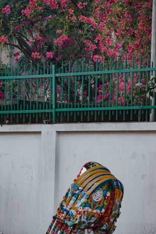 A richly colored textile displays intricate designs with a variety of patterns, fruits, and floral motifs. It is set against a plain white wall backdrop, with vibrant pink flowers and lush green foliage in the background, partially obscured by a metal fence.