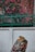 A richly colored textile displays intricate designs with a variety of patterns, fruits, and floral motifs. It is set against a plain white wall backdrop, with vibrant pink flowers and lush green foliage in the background, partially obscured by a metal fence.
