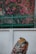 A richly colored textile displays intricate designs with a variety of patterns, fruits, and floral motifs. It is set against a plain white wall backdrop, with vibrant pink flowers and lush green foliage in the background, partially obscured by a metal fence.