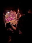 A person is sitting in front of a small shrine or altar, reading a religious or spiritual book. The setting is dimly lit with a soft, warm glow highlighting religious icons, flowers, and offerings. The scene exudes a sense of devotion and calmness.