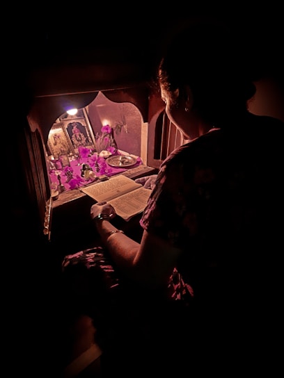 A person is sitting in front of a small shrine or altar, reading a religious or spiritual book. The setting is dimly lit with a soft, warm glow highlighting religious icons, flowers, and offerings. The scene exudes a sense of devotion and calmness.