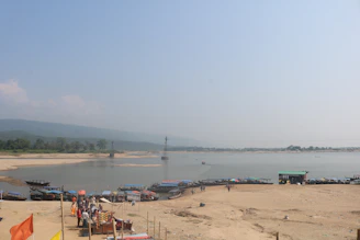A riverside area with a sandy shore and several small boats parked along the edge of the water. People are gathered near the boats, some engaged in various activities. In the background, hills and sparse vegetation can be seen under a clear, blue sky.