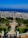 A beautifully landscaped garden with symmetrical design, featuring a central staircase leading down towards lush greenery. Palm trees and ornate hedges line the path, adding an exotic touch. The view extends to a bustling city below, with a vibrant blue sea and clear sky in the background.