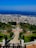 A beautifully landscaped garden with symmetrical design, featuring a central staircase leading down towards lush greenery. Palm trees and ornate hedges line the path, adding an exotic touch. The view extends to a bustling city below, with a vibrant blue sea and clear sky in the background.