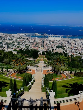 A beautifully landscaped garden with symmetrical design, featuring a central staircase leading down towards lush greenery. Palm trees and ornate hedges line the path, adding an exotic touch. The view extends to a bustling city below, with a vibrant blue sea and clear sky in the background.