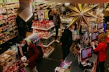 A busy store with several people wearing masks, shopping and checking out goods. Colorful shelves are stocked with a variety of items, including candy and snacks. A large decorative star hangs prominently above the checkout area, where a cashier assists a customer at the register.