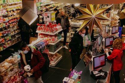 A busy store with several people wearing masks, shopping and checking out goods. Colorful shelves are stocked with a variety of items, including candy and snacks. A large decorative star hangs prominently above the checkout area, where a cashier assists a customer at the register.
