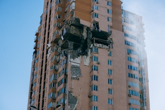 A tall residential building with significant structural damage on one side, exposing the interior and causing debris to hang precariously. The blue sky provides a stark contrast to the damaged structure.