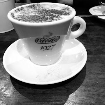 A white ceramic coffee cup filled with a frothy beverage, placed on a matching white saucer. The cup has the branding 'Carraro 1927' on it. The background includes a wooden table surface and a white plate with a spoon in the top right corner.