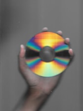 A hand holding a compact disc, which displays a spectrum of colors ranging from bright red, through orange, yellow, green, and blue, against a gray background.